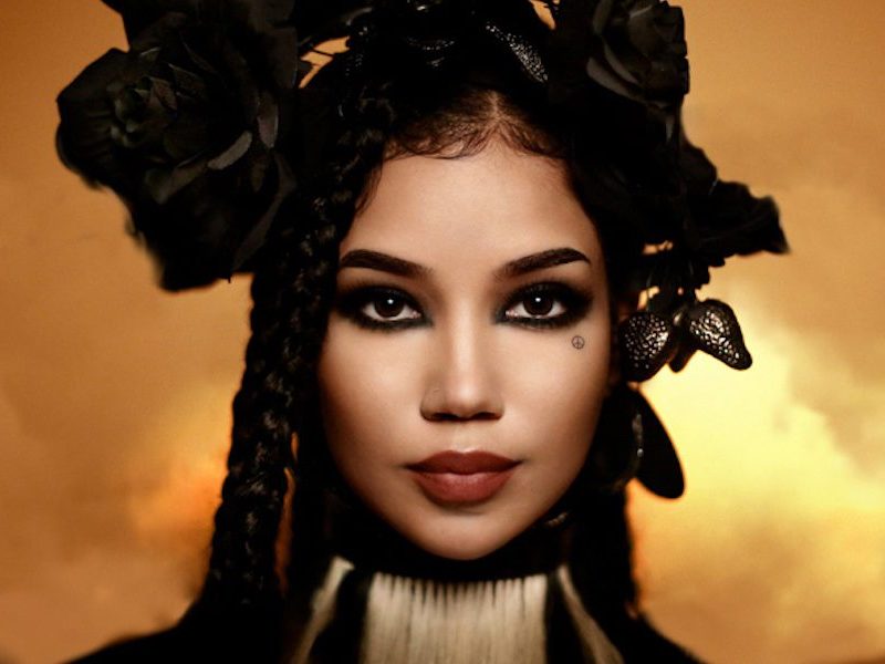 Photo of Jhene Aiko for Chilombo deluxe album. She has box braids and black flowers in her hair; her makeup is dark and heavy.